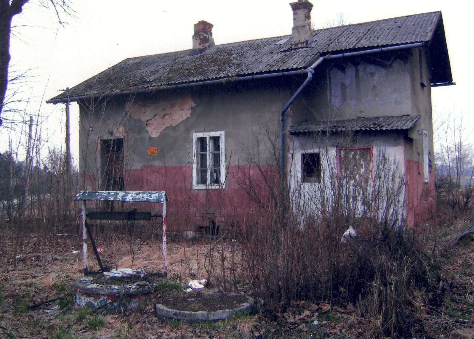 Opuszczony parterowy dom z odpadającym tynkiem i eternitowym dachem, przed nim studnia, wokół bezlistne krzewy.