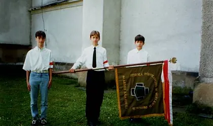 Trójka uczniów w strojach galowych ze sztandarem szkoły przed białą ścianą.