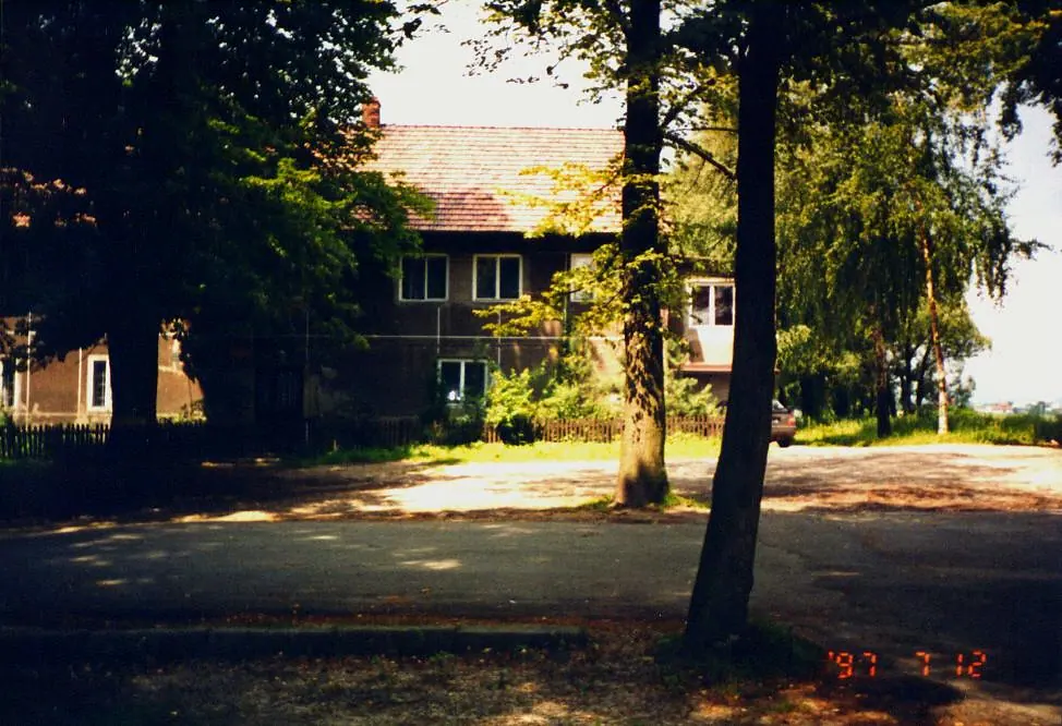 Parterowy budynek wikarówki wśród wysokich drzew.