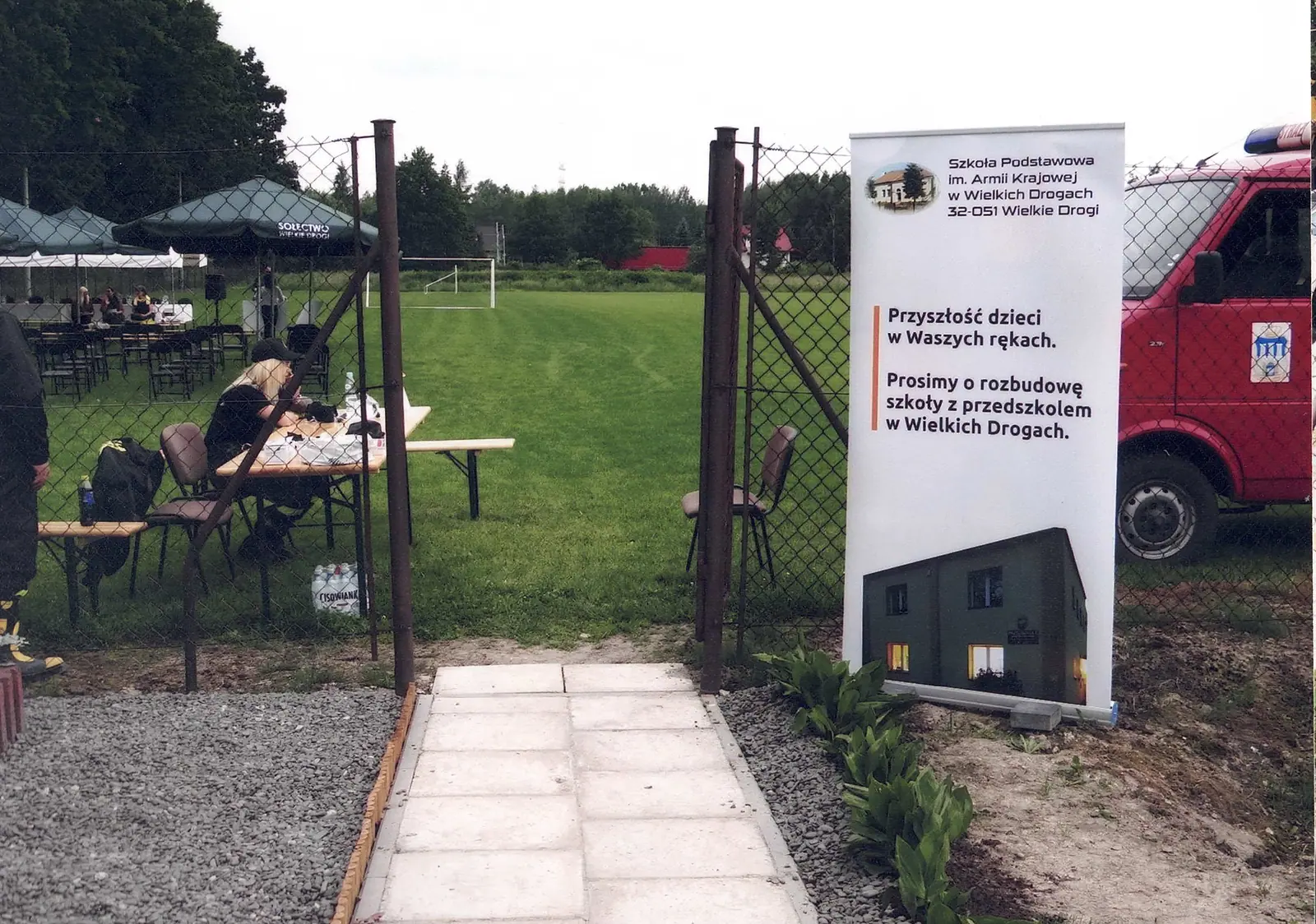 Wejście na boisko sportowe przez bramę w siatce. Przy bramie stoi baner reklamowy z apelem o rozbudowę szkoły, obok wóz strażacki, w głębi stoły i namioty przygotowane na spotkanie.