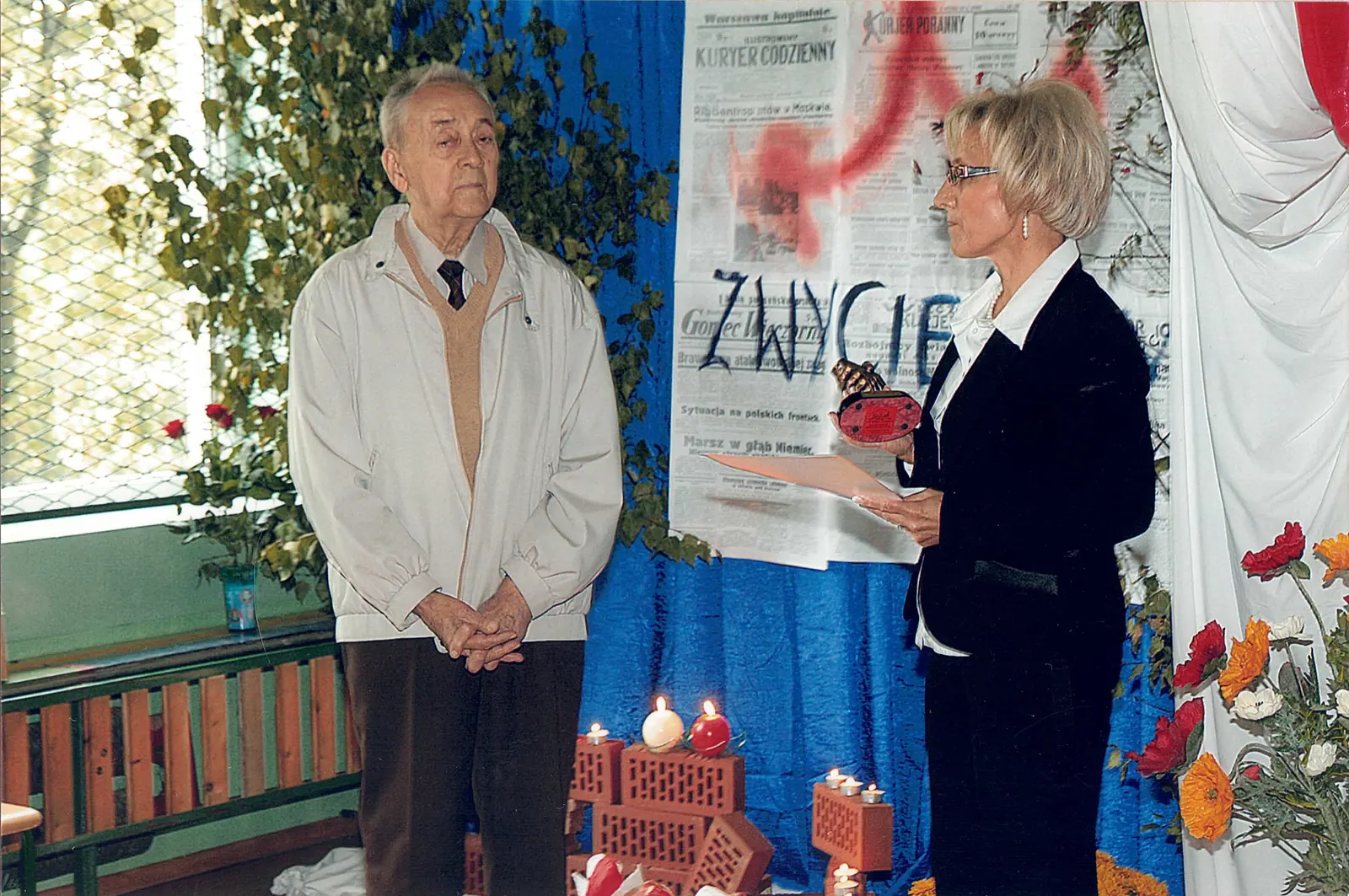 Starszy mężczyzna w jasnej kurtce stoi ze złożonymi dłońmi, naprzeciw kobieta odczytuje dyplom i trzyma statuetkę. W tle dekoracja z powiększonych pierwszych stron gazet z września 1939 roku, biało-czerwona tkanina i znicze.