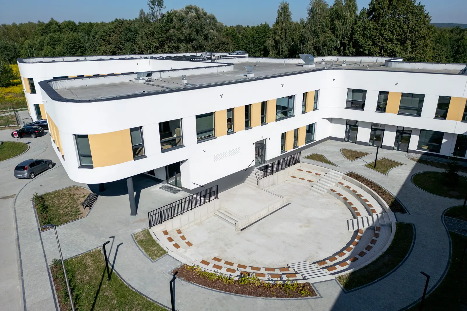 Półkolisty amfiteatr z betonowymi stopniami i drewnianymi siedziskami, wokół niego zakrzywione skrzydło szkoły wsparte na słupach.