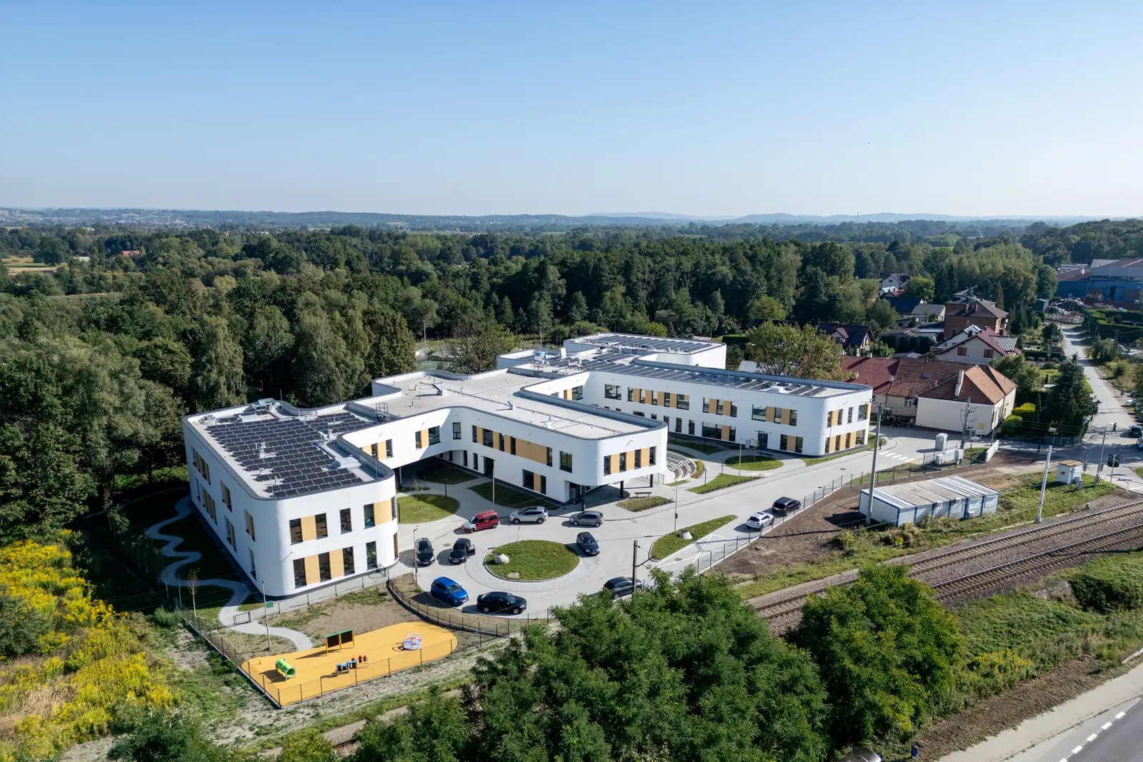 Cały kompleks z lotu ptaka, na dachach panele słoneczne, po lewej plac zabaw, w tle lasy i pola.
