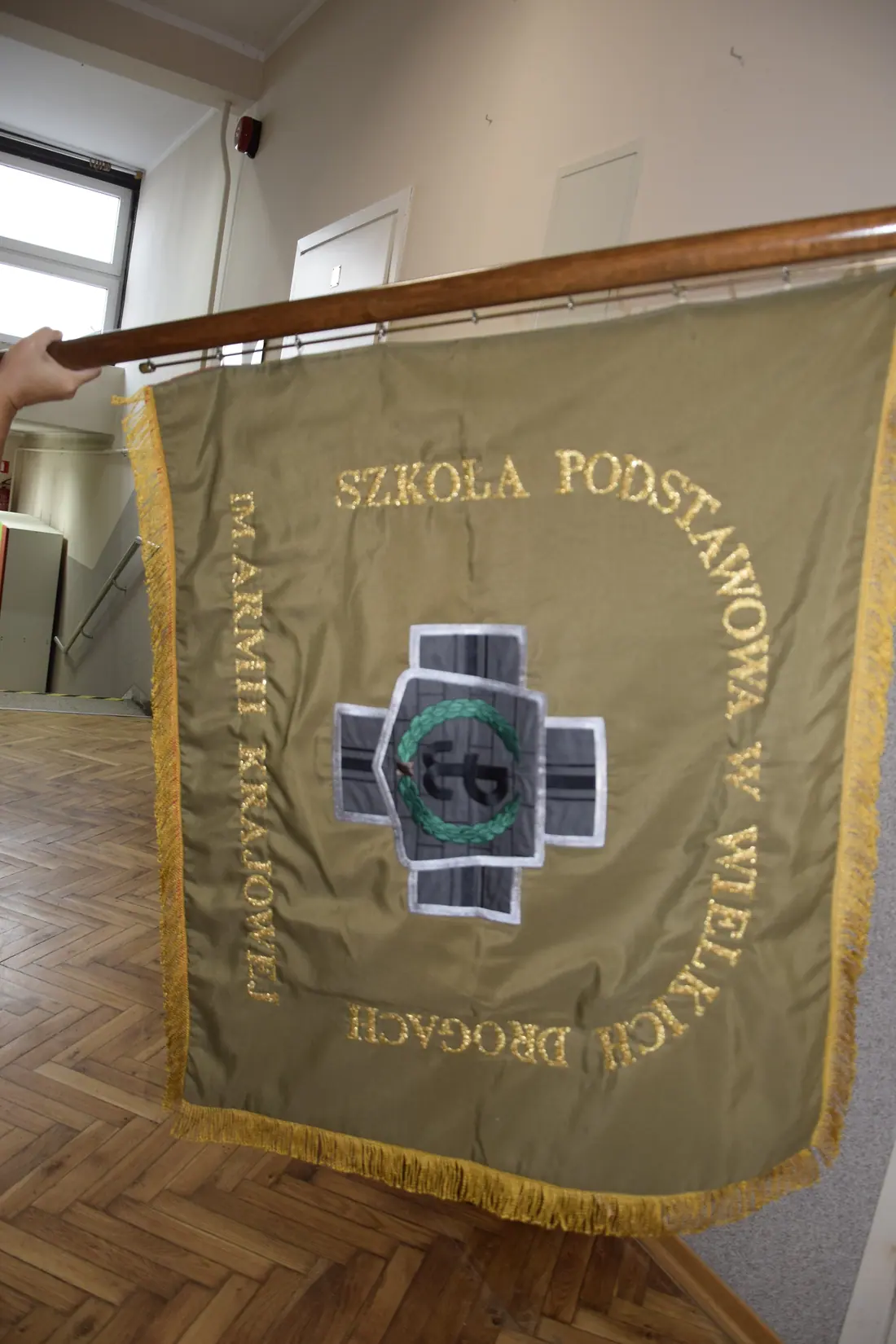 Sztandar na drzewcu: oliwkowy płat ze złotymi frędzlami, pośrodku haftowany Krzyż Armii Krajowej, dokoła złoty napis z nazwą szkoły.