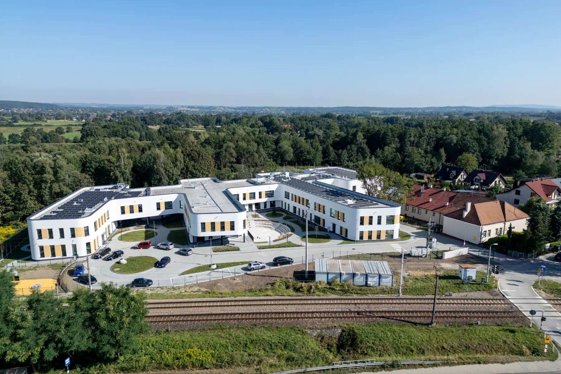 Gotowy kompleks Centrum Edukacyjnego w słońcu, samochody na parkingu, wzdłuż działki tory kolejowe, w tle zabudowa wsi.