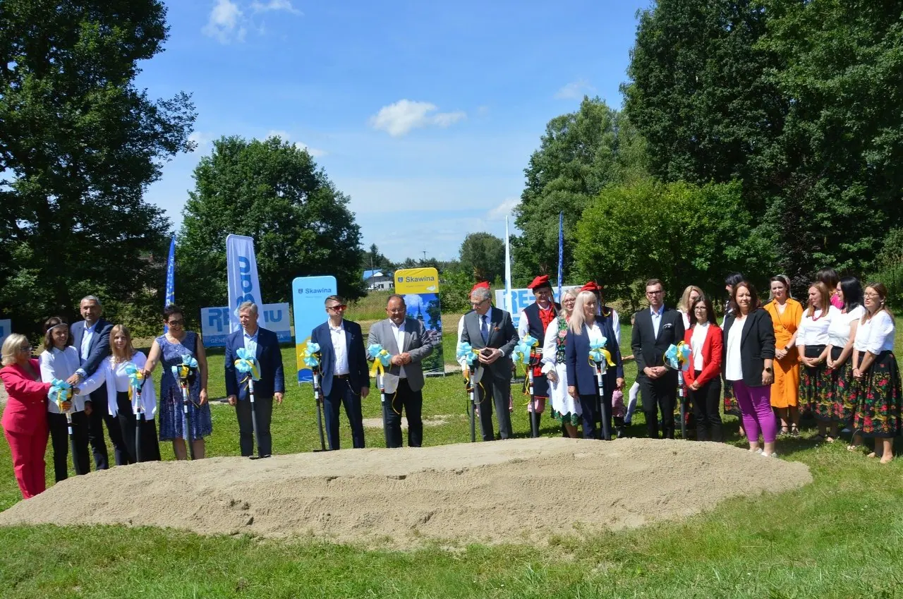 Kilkanaście osób stoi w szeregu za kopcem piasku, każda trzyma łopatę z niebiesko-żółtą kokardą. W tle banery Gminy Skawina i RE-Bau.