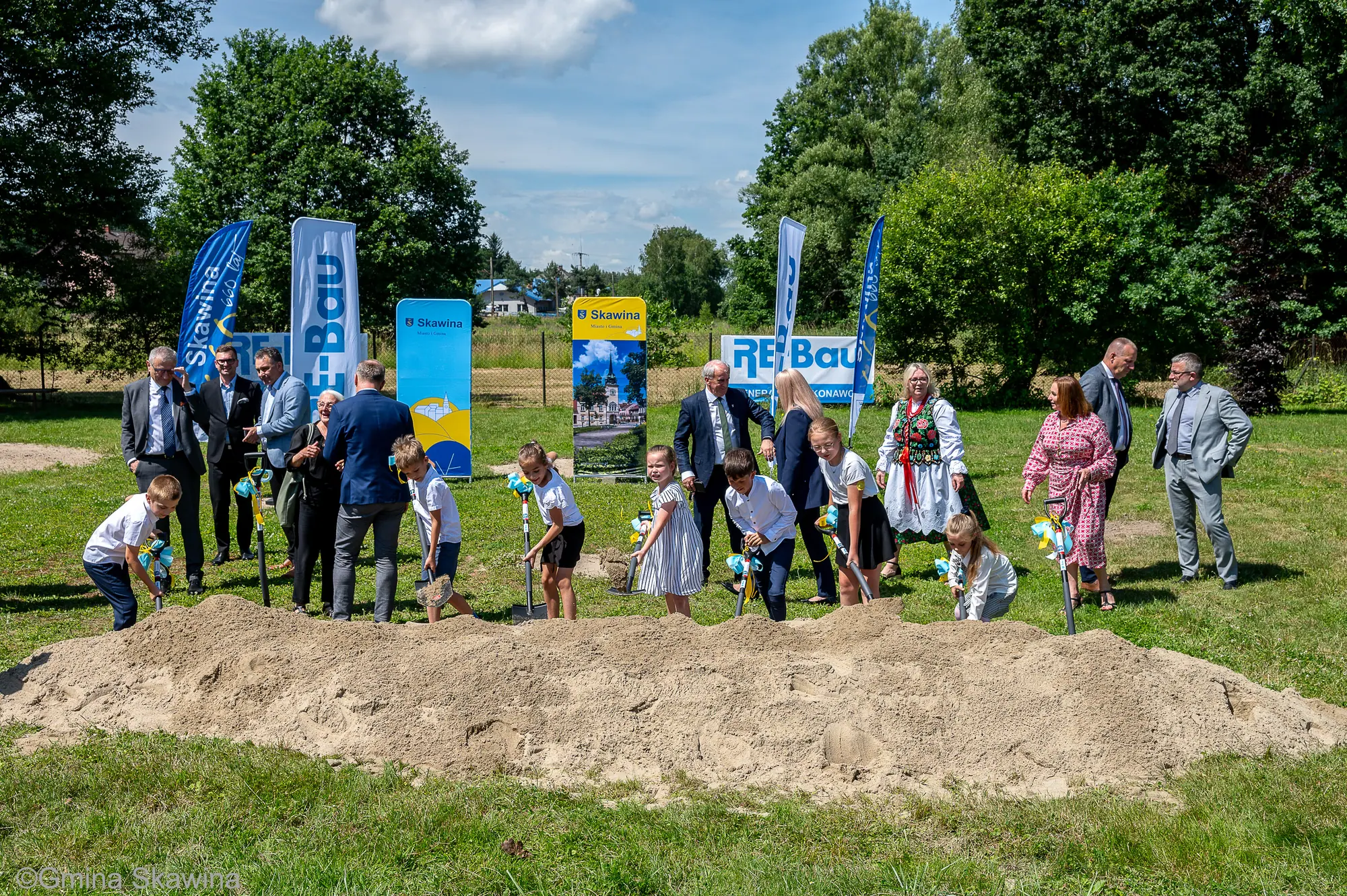 Grupa osób z łopatami wokół kopca ziemi na placu przyszłej budowy.