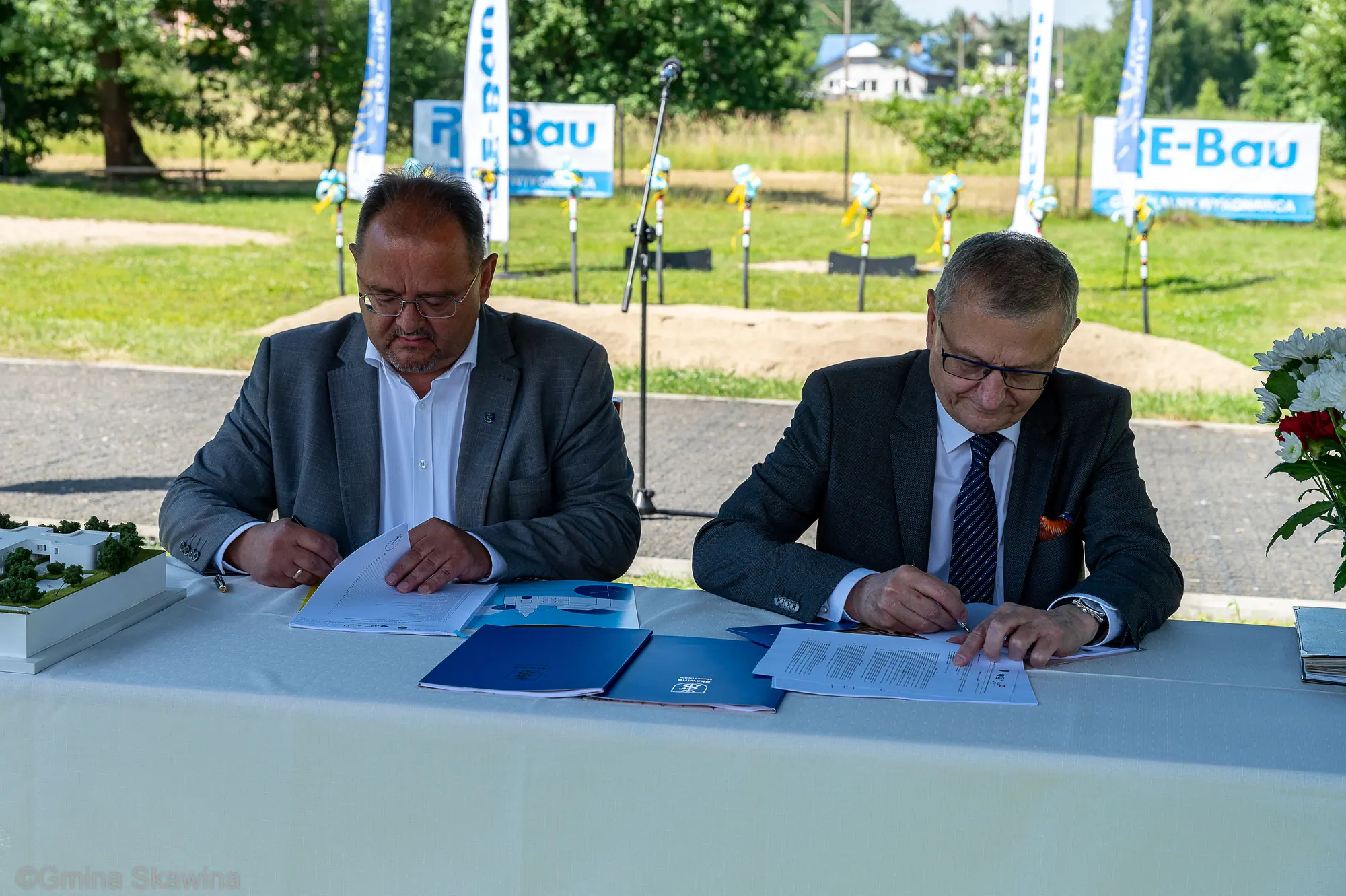 Dwaj mężczyźni podpisują dokumenty przy stole nakrytym białym obrusem.