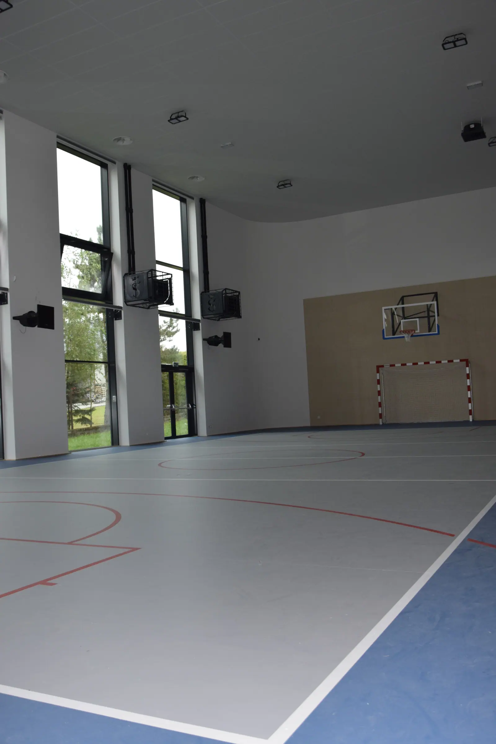 Wnętrze sali gimnastycznej z jasną i niebieską wykładziną sportową, drabinkami przy ścianie i bramką do piłki ręcznej.