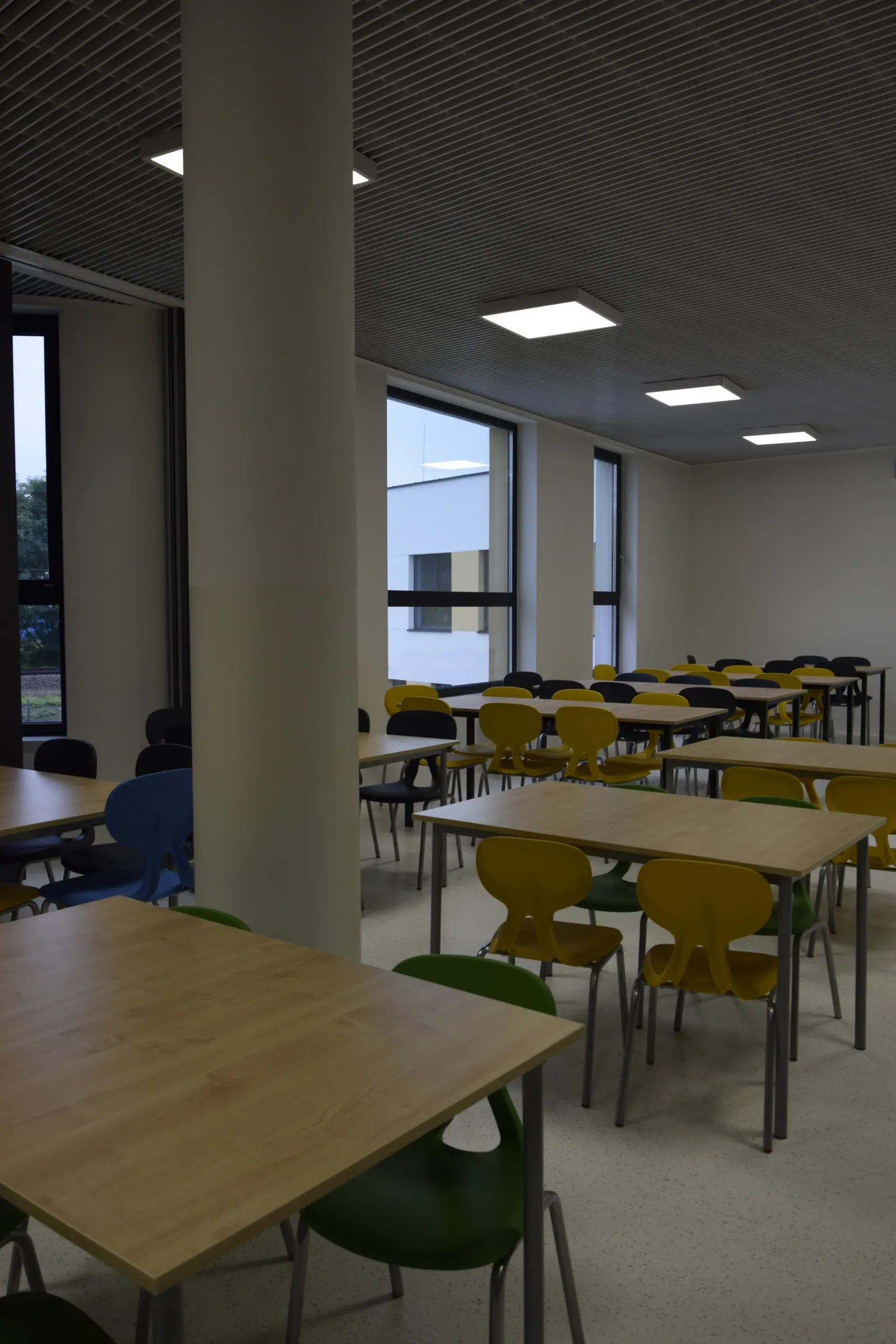 Jadalnia z drewnianymi stołami i krzesłami w kolorach żółtym i zielonym, przy oknie sięgającym podłogi.