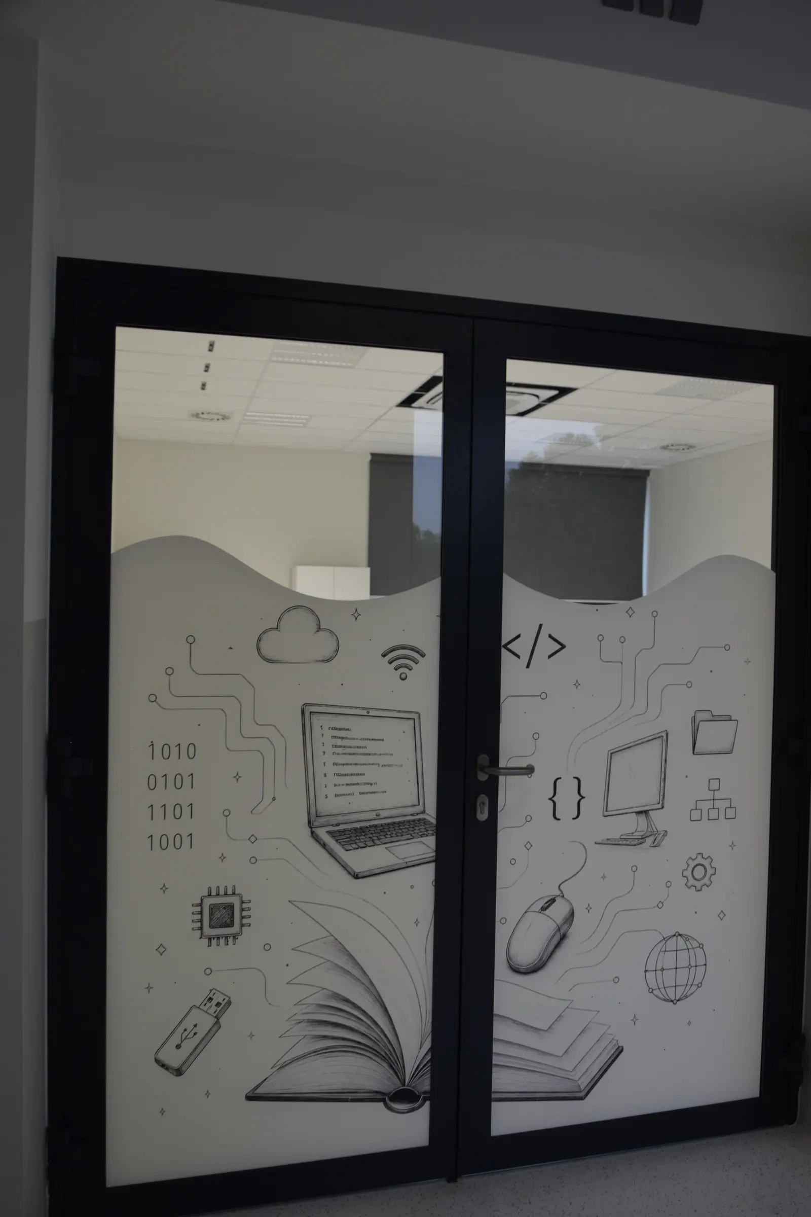 Przeszklona ściana pracowni komputerowej z matową grafiką: laptop, chmura, znaczniki kodu i obwody drukowane.