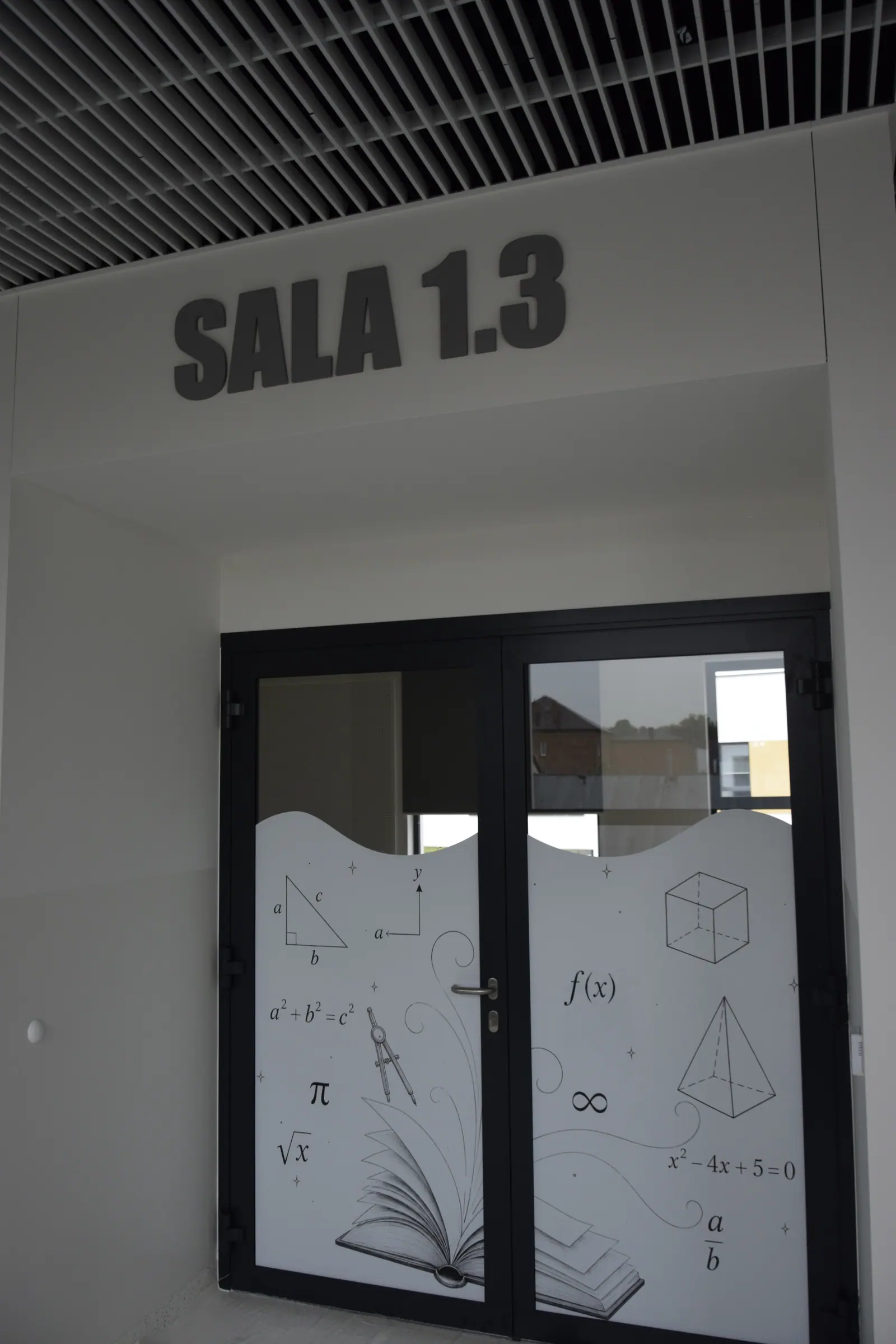 Przeszklone drzwi sali lekcyjnej z matową grafiką przedstawiającą wzory i bryły geometryczne, nad nimi napis SALA 1.3.